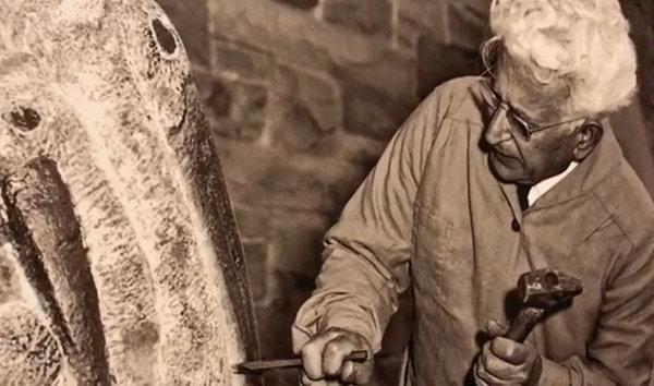 Black and white image of Henry Dispirito at work on a sculpture, with hammer and chisel in hand.